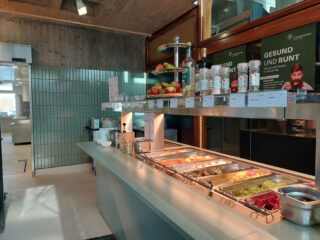 Eine Selbstbedienungstheke in einer Cafeteria mit Tabletts voller Essen, Äpfeln, Gewürzen und Schildern, die auf Deutsch für gesunde Ernährung werben. Im Hintergrund sind eine geflieste Wand und ein Küchenbereich zu sehen.