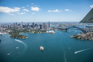 Luftaufnahme des Hafens von Sydney mit dem Sydney Opera House, der Sydney Harbour Bridge, der Skyline der Innenstadt und Booten im blauen Wasser an einem sonnigen Tag.