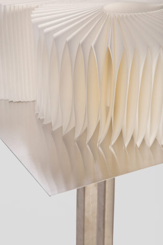 Eine Nahaufnahme einer geometrischen Papierskulptur mit gleichmäßig verteilten, gefalteten Segmenten auf einer reflektierenden Metalloberfläche vor einem unifarbenen Hintergrund.