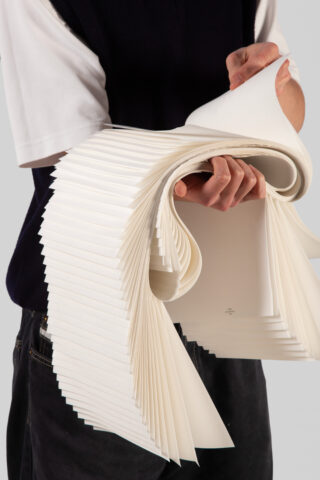 Eine Person hält mit beiden Händen einen übergroßen Stapel weißer, gebogener und aufgefächerter Papierblätter vor einem unifarbenen Hintergrund.