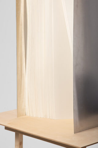 Nahaufnahme eines großen Stapels weißer Blätter, teilweise umrahmt von einem Metallblatt auf der rechten Seite, das aufrecht auf einem hellen Holztisch steht.