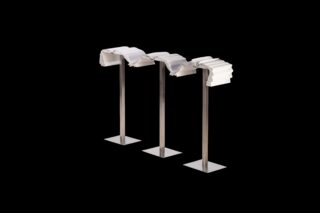 Drei Metallständer tragen jeweils einen Stapel weißer, gefalteter Papiere, die vor einem schwarzen Hintergrund drapiert sind.