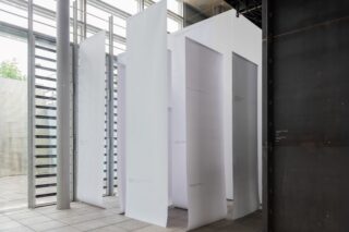 In einem modernen Raum im Industriestil mit großen Fenstern hängen hohe, weiße vertikale Paneele von der Decke und bilden eine labyrinthartige Installation.