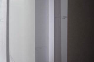 Weiße Papierbögen mit schwachem, klein gedrucktem Text hängen vertikal in parallelen Reihen und bilden eine minimalistische und abstrakte Komposition.