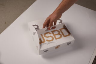 Eine Hand hebt eine weiße Pappschachtel mit braunen Buchstaben und kleinen Illustrationen an, die auf einer weißen Fläche steht.