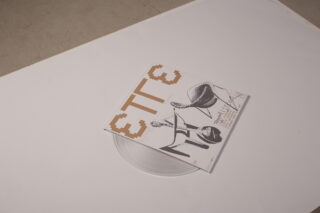 Eine durchsichtige Vinyl-Schallplatte mit einem Cover mit abstraktem schwarz-weißem Artwork und fettgedrucktem braunem Text wird auf eine weiße Oberfläche gelegt.