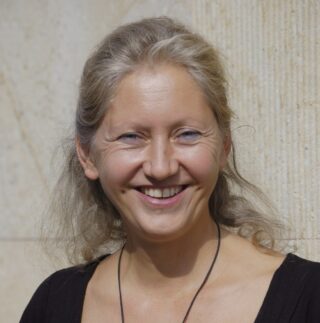 Eine Frau mit hellbraunem Haar lächelt in die Kamera, während sie vor einer beigefarbenen, strukturierten Wand steht. Sie trägt ein schwarzes Oberteil und eine Halskette.