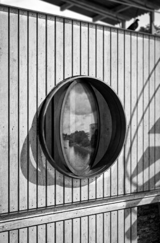 Ein rundes Fenster im Stil eines Bullauges, das in eine Holzlattenwand eingelassen ist und eine Szene am Wasser widerspiegelt; auf dem Dach darüber sitzt ein Vogel.