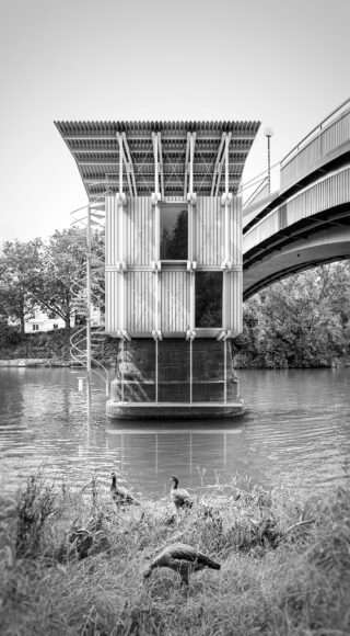 Ein modernes Gebäude mit einer Wendeltreppe steht an einem Fluss, neben einer Brücke; am grasbewachsenen Flussufer im Vordergrund sind Enten zu sehen.