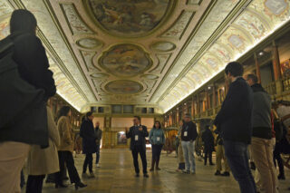 Eine Gruppe von Menschen hört einem Fremdenführer in einer verzierten Halle mit verzierten Decken und Kunstwerken an den Wänden zu.