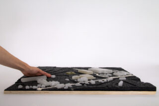 Eine Hand berührt ein geschichtetes architektonisches Geländemodell mit weißen Gebäudeformen und grauem Terrain auf einem hölzernen Sockel vor einem schlichten weißen Hintergrund.