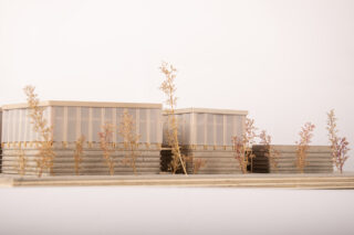 Ein maßstabsgetreues Architekturmodell zeigt zwei rechteckige Gebäude mit gestapelten Sockeln und durchsichtigen oberen Wänden, umgeben von kleinen Modellbäumen auf einer flachen Plattform.