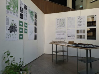 Eine Ausstellung mit architektonischen Zeichnungen und Diagrammen an weißen Wänden, im Vordergrund ein Tisch mit Architekturmodellen und Materialien. Kleine Pflanzen stehen auf dem Boden.