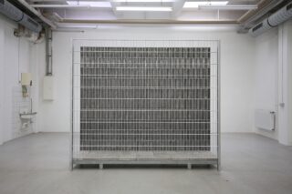 Ein großer Metallkäfig steht in der Mitte eines kargen, industriell anmutenden weißen Raums mit sichtbaren Rohren und einem an der Wand befestigten Waschbecken.