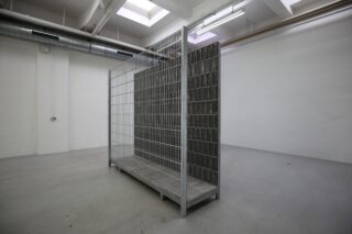 In einem leeren, weißwandigen Raum mit Betonboden und freiliegenden Deckenrohren steht eine Käfigkonstruktion aus Metalldraht mit einer massiven dunklen Platte im Inneren.