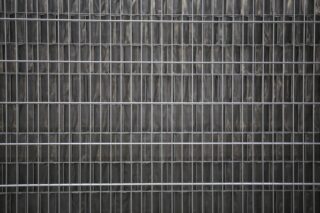 Eine Nahaufnahme eines Metalldrahtzauns mit vertikalen und horizontalen Stäben vor einem dunklen, strukturierten Hintergrund.