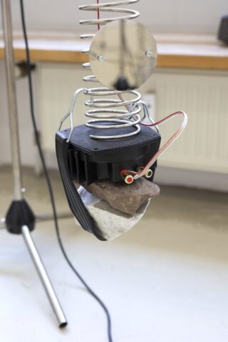 Nahaufnahme einer abstrakten Skulptur mit einer gewundenen Metallfeder, einem runden Spiegel, Drähten, Steinen und einem schwarzen Kunststoffteil, die auf einem beigen Boden steht.