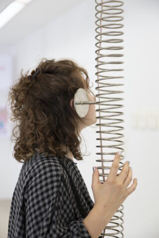 Eine Person mit lockigem Haar hört eine große Metallspule und drückt ein Ohr an eine kreisförmige Platte, die an der Spule in einem weißen Raum befestigt ist.