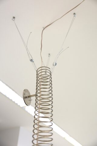 Eine große Metallfeder wird senkrecht an der Wand montiert, wobei dünne Drähte von der Spitze ausgehen und an der Decke befestigt werden.