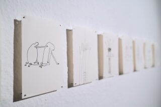 Eine Reihe minimalistischer Strichzeichnungen auf weißem Papier ist in einer Kunstgalerie an eine weiße Wand gepinnt.