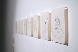 Eine Reihe von minimalistischen Strichzeichnungen auf weißem Papier ist an eine weiße Wand in einer Kunstgalerie gepinnt.
