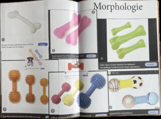 Eine Katalogseite zeigt verschiedene bunte Kauspielzeuge für Hunde in Form von Knochen, Hanteln und Bällen vor einem weißen Hintergrund mit Produktnamen und Preisen in deutscher Sprache.