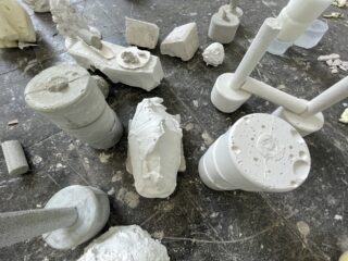 Verschiedene weiße Gipsskulpturen und geometrische Formen sind auf einem rauen, dunklen Boden angeordnet.