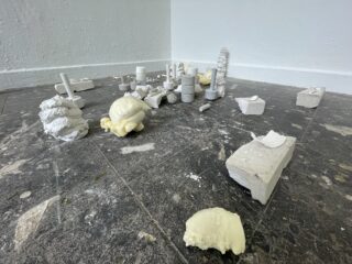 In einem galerieähnlichen Raum sind verschiedene Gips- und Schaumstoffskulpturen auf einem abgewetzten, dunklen Kachelboden vor einer weißen Wand angeordnet.