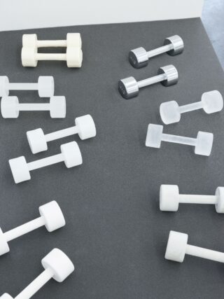 Eine Vielzahl von Hanteln, meist weiß und eine aus Metall, sind auf einer dunklen Fläche vor einer weißen Wand angeordnet.