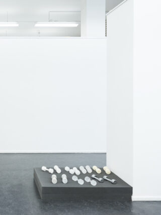 Auf einem schwarzen Podest sind in einem minimalistischen, weißwandigen Raum mehrere kleine, identische Objekte in Reihen angeordnet.