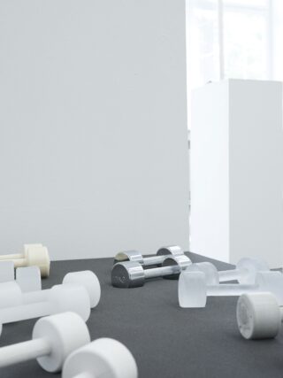 Mehrere helle Hanteln und eine Metallhantel werden auf einer dunklen Fläche in der Nähe einer weißen Wand in einem minimalistischen, gut beleuchteten Raum platziert.