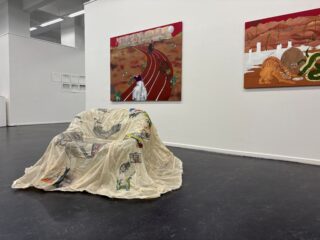 In einer Galerie steht ein mit Stoff bespannter Stuhl mit bunten Skizzen, an der weißen Wand dahinter hängen zwei große, lebhaft gemalte Kunstwerke.