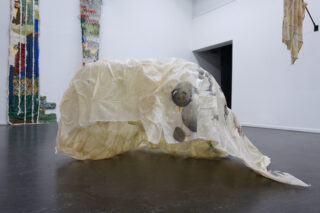 Eine durchsichtige Plastikfolie, die über ein Objekt auf dem Boden einer Galerie drapiert ist, wobei eine Bärenillustration auf der Folie zu sehen ist; an den Wänden im Hintergrund hängen textile Kunstwerke.