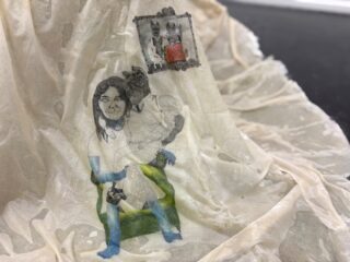 Ein durchsichtiger, drapierter Stoff zeigt eine Zeichnung von zwei Menschen und einem Wolf, mit einem gerahmten Bild eines Wolfes über ihnen. Das Kunstwerk ist mit schwarzen, grünen, blauen und roten Akzenten versehen.