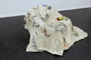 Ein weißer Sessel ist mit einem durchsichtigen, illustrierten Stoff bezogen, auf dem verschiedene Skizzen und farbige Zeichnungen zu sehen sind, und steht in einem schlichten Raum mit dunklem Fußboden.