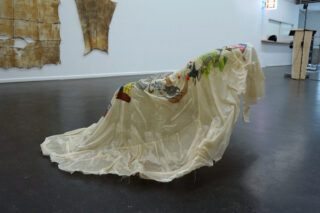 Ein lichtdurchlässiger, dekorierter Stoff wird über eine gewinkelte Struktur in einem Galerieraum mit Kunstwerken an den Wänden und einem Podium im Hintergrund drapiert.