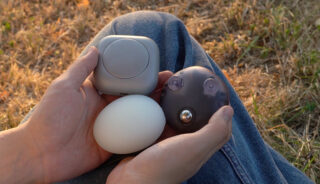 Zwei Hände halten drei kleine, runde elektronische Geräte - ein silbernes, ein weißes, eiförmiges und ein dunkles mit kleinen Ausbuchtungen - über den mit Jeans bekleideten Knien im Freien auf trockenem Gras.