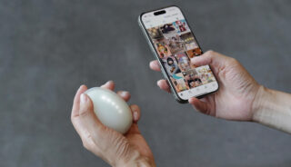 Eine Person hält in der einen Hand ein weißes Ei und in der anderen Hand ein Smartphone, das vor einem grauen Hintergrund einen Social-Media-Feed anzeigt.