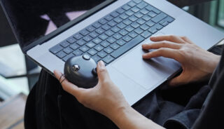 Eine Person benutzt einen Laptop mit beiden Händen, eine Hand auf der Tastatur und die andere hält eine kabelgebundene Trackball-Maus.