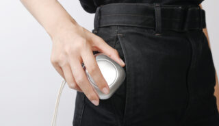 Eine Person steckt ein kleines, silbernes medizinisches Gerät, das an einem Schlauch befestigt ist, in die Gesäßtasche einer schwarzen Jeans.