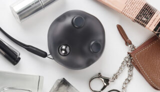 Ein runder schwarzer Gegenstand mit Knöpfen und einer Metallkugel wird auf einer weißen Fläche neben Schminke, einem Schlüsselbund und anderen persönlichen Gegenständen gezeigt.