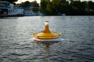 Eine gelbe schwimmende Boje befindet sich auf einem ruhigen Gewässer, in dessen Hintergrund Bäume und Boote zu sehen sind.