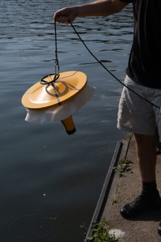 Eine Person hält ein gelbes, scheibenförmiges schwimmendes Gerät, das an einem Kabel über dem Rand eines Gewässers befestigt ist.