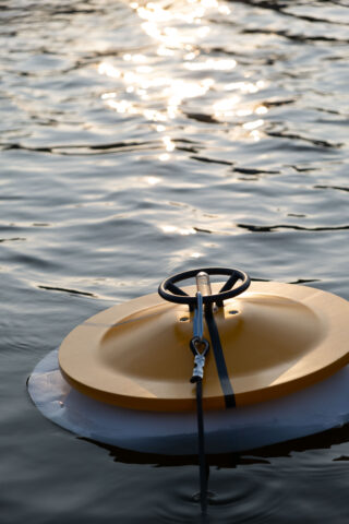 Ein gelbes, kreisförmiges Objekt schwimmt auf dem Wasser, angebunden an einer schwarzen Schnur, und das Sonnenlicht spiegelt sich auf der kräuselnden Oberfläche.