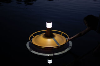Eine Person setzt ein schwimmendes, gelbes Gerät mit einer brennenden Lampe an der Spitze in dunkles Wasser.
