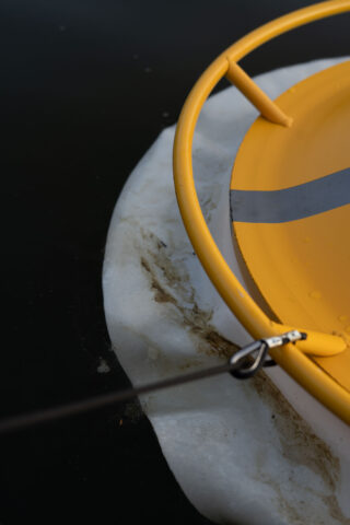Nahaufnahme einer gelben Rettungsboje mit einem grauen Streifen, die mit einer Metallklammer und einem Seil auf einem Stück Eis ruht, vor einem dunklen Hintergrund.