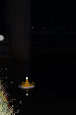Eine gelbe Boje schwimmt nachts im dunklen Wasser in der Nähe einer Betonbrücke, im Hintergrund sind entfernte Straßenlaternen zu sehen.