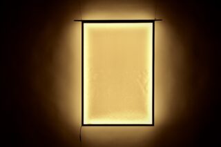 Rechteckiges Lichtpaneel mit leuchtend gelbem Farbton, das an einer Wand montiert ist und ein warmes, weiches Licht abgibt. An der Unterkante des Rahmens hängt ein Stromkabel herunter.