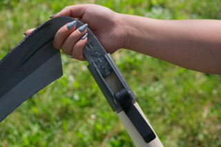 Die Hand einer Person befestigt ein Sensenblatt aus Metall an einem Holzgriff im Freien auf einem grasbewachsenen Hintergrund.