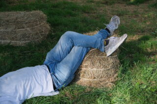 Person im Gras liegend, die Beine auf einen Heuballen gestützt, in blauen Jeans, weißem Hemd und weißen Turnschuhen. Ein weiterer Heuballen ist in der Nähe zu sehen.